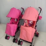 Dos cochecitos tipo sombrilla en color rosa, con capota protectora y cestas frontales, sobre suelo de baldosas blancas.