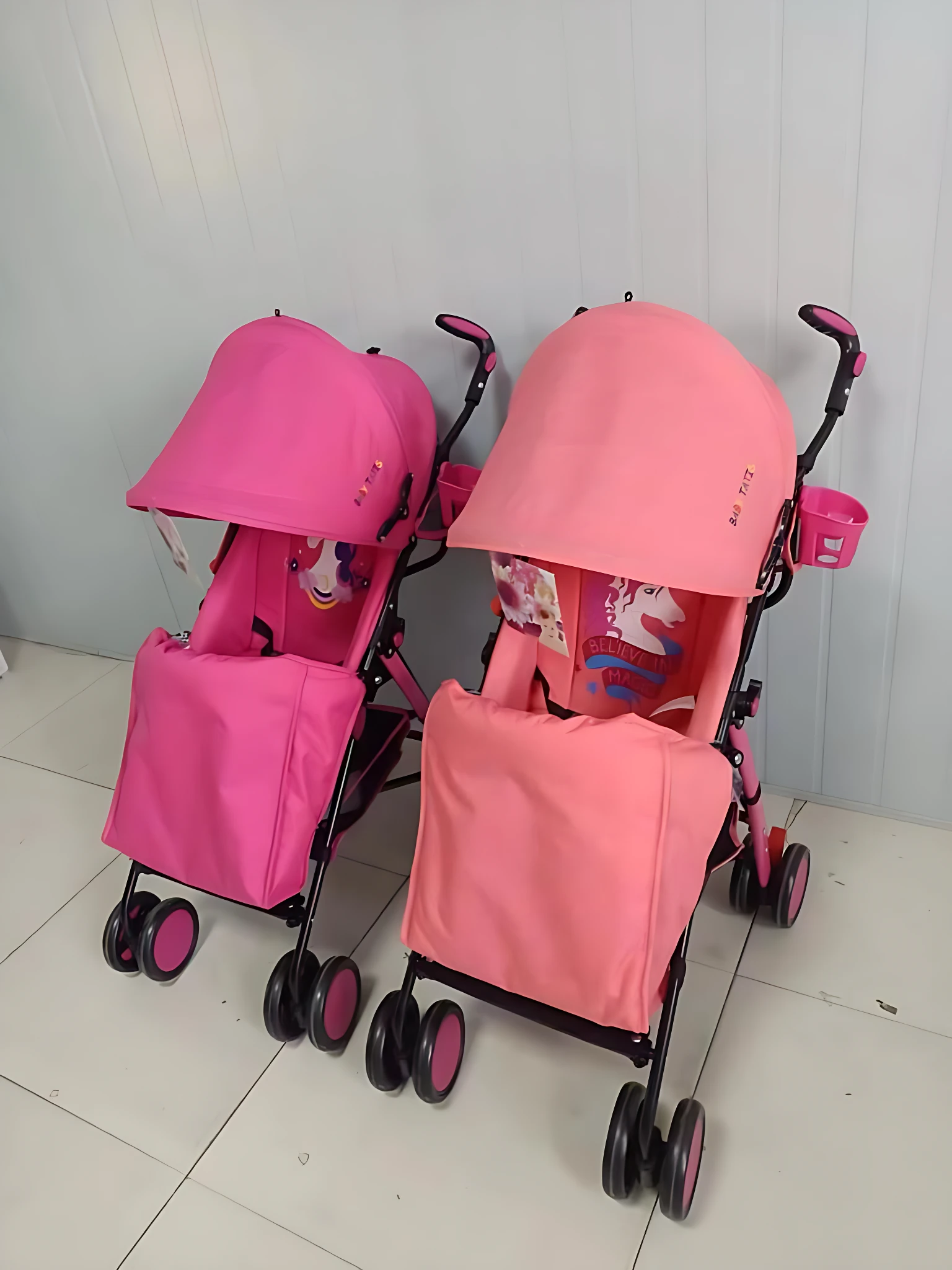 Dos cochecitos tipo sombrilla en color rosa, con capota protectora y cestas frontales, sobre suelo de baldosas blancas.