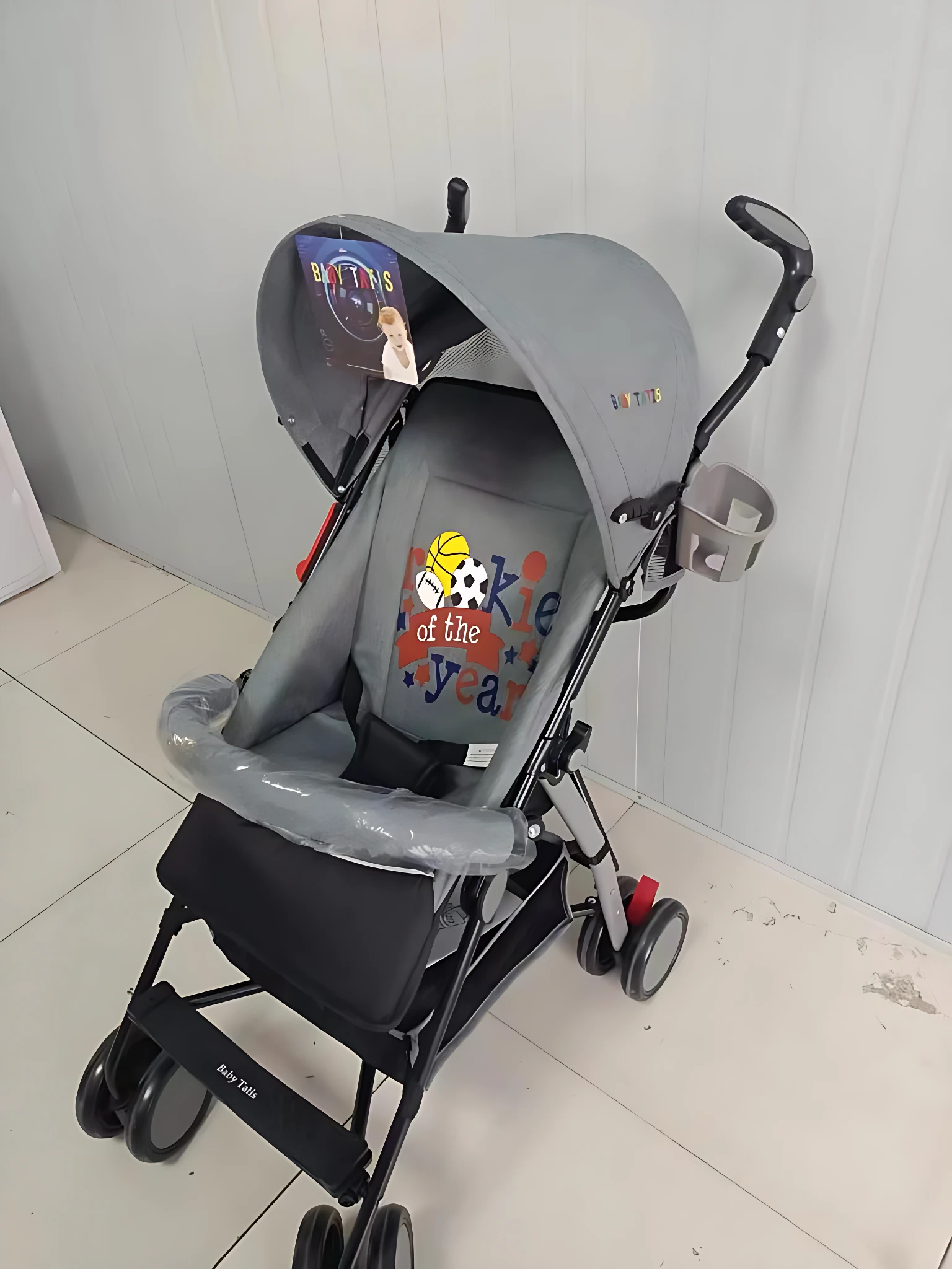 Coche paseador tipo sombrilla en gris con capota protectora, asiento acolchado y detalles gráficos infantiles. Incluye reposapiés plegable y ruedas pequeñas para desplazamiento fácil.