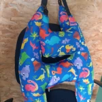 Cojín de viaje en forma de H con estampado de dinosaurios multicolores sobre fondo azul, colocado sobre asiento de silla.