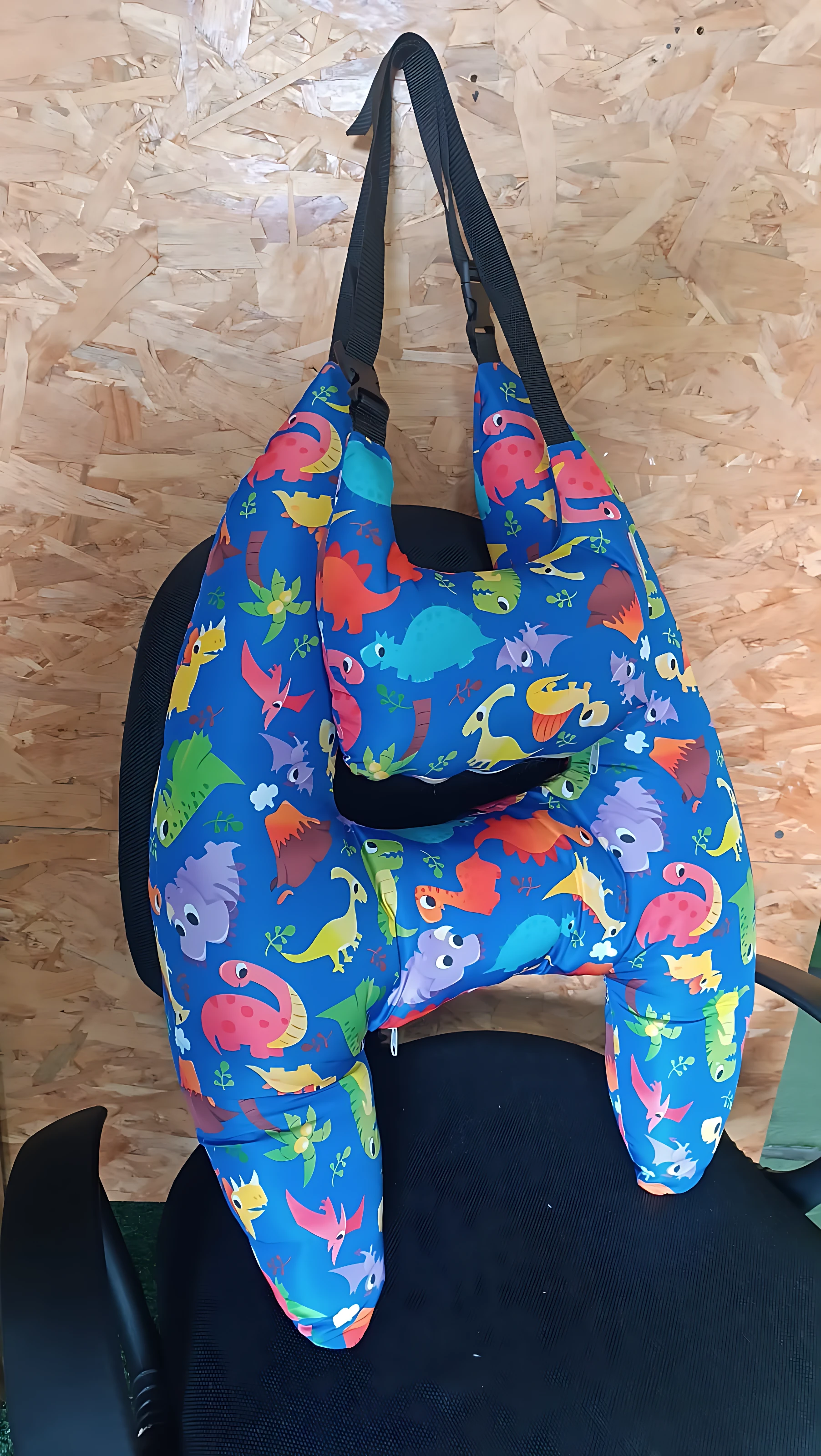 Cojín de viaje en forma de H con estampado de dinosaurios multicolores sobre fondo azul, colocado sobre asiento de silla.