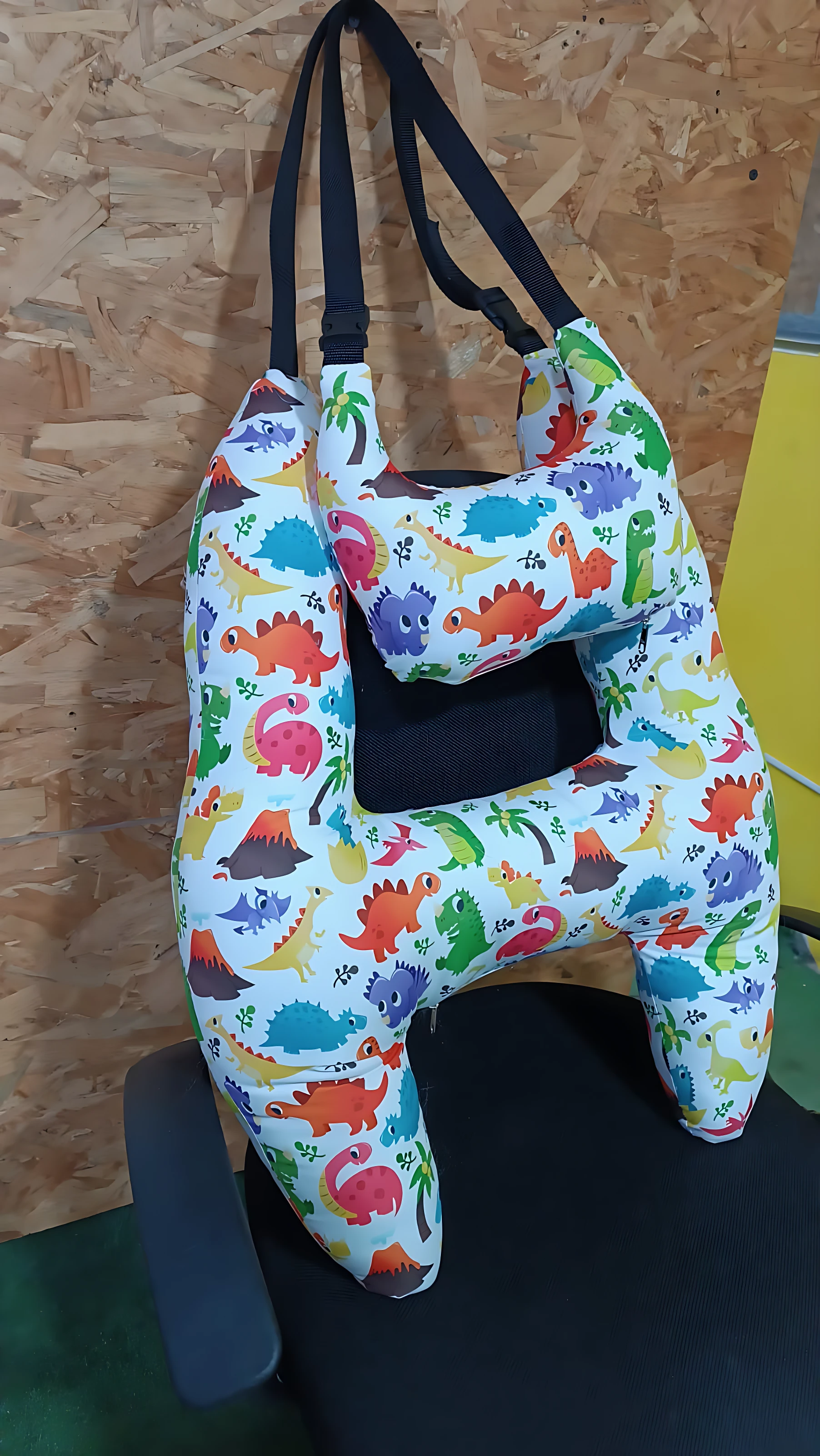 Cojín de viaje en forma de H con estampado de dinosaurios multicolores sobre fondo blanco, mostrando su estructura ergonómica y correas negras ajustables.