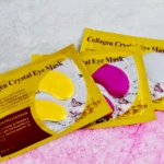 Tres paquetes de parches oculares en colores amarillo, magenta y blanco, sobre fondo rosa texturizado. Cada uno muestra dos almohadillas transparentes con forma de ojo.