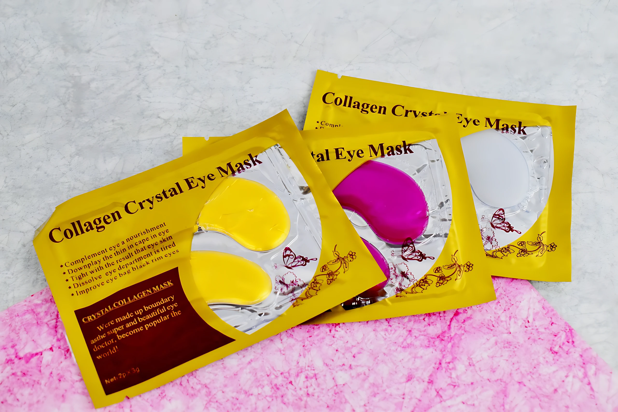 Tres paquetes de parches oculares en colores amarillo, magenta y blanco, sobre fondo rosa texturizado. Cada uno muestra dos almohadillas transparentes con forma de ojo.