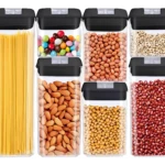 Siete contenedores herméticos de plástico transparente con tapas negras, mostrando pasta, almendras, cacahuetes, legumbres y dulces.