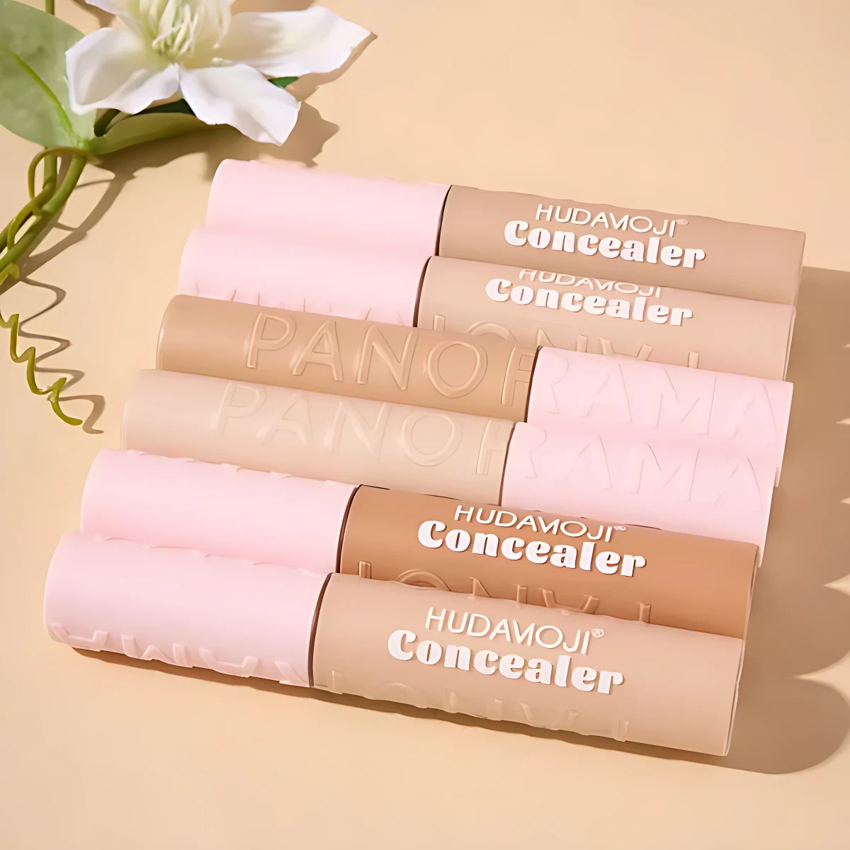 Set de dos correctores Hudamoji en tonos nude y rosa, con acabado mate y empaque cilíndrico, dispuestos diagonalmente sobre fondo beige claro junto a una flor blanca.