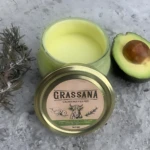 Tapa de crema corporal natural de aguacate y romero, con etiqueta que muestra el nombre del producto y una de un animal. Al lado, una rodaja de aguacate y una rama de romero sobre superficie gris.