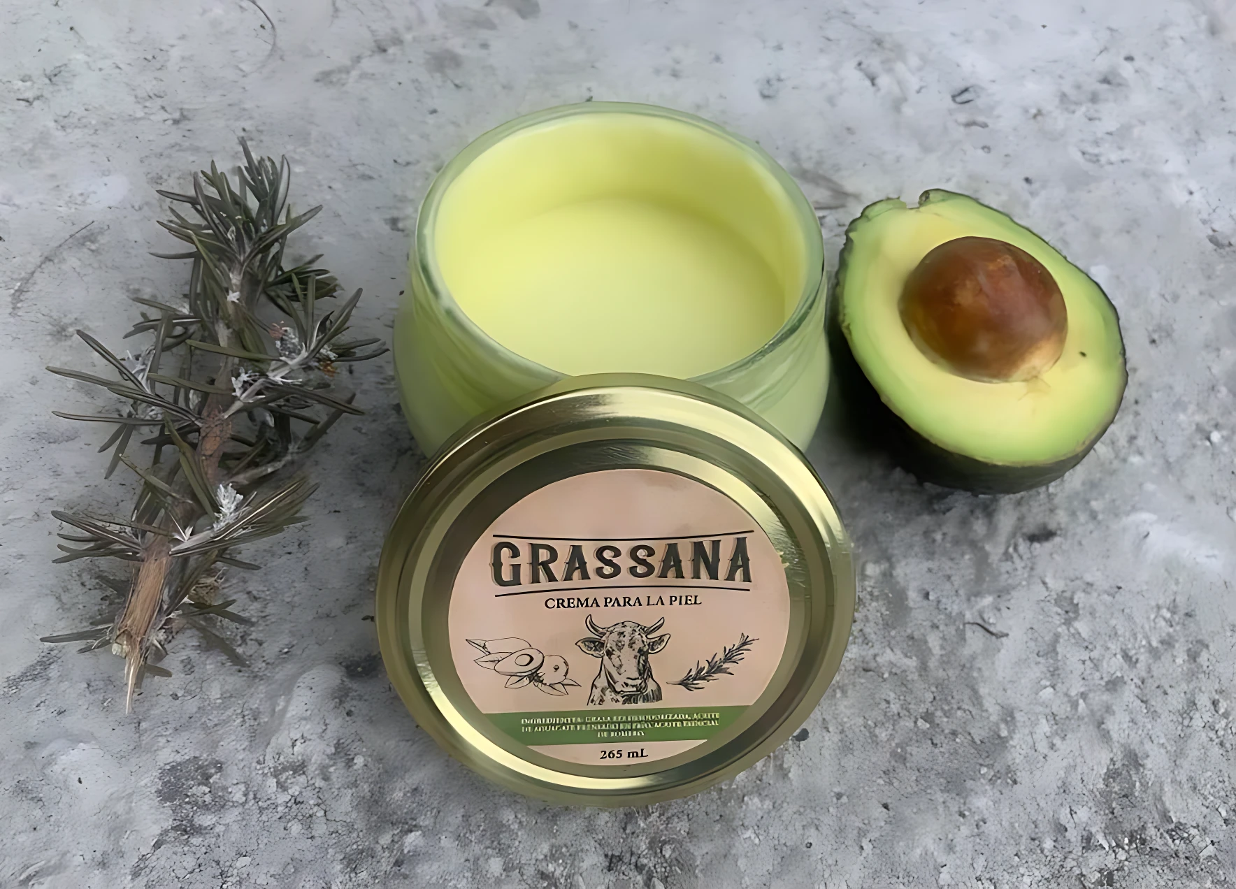 Tapa de crema corporal natural de aguacate y romero, con etiqueta que muestra el nombre del producto y una de un animal. Al lado, una rodaja de aguacate y una rama de romero sobre superficie gris.