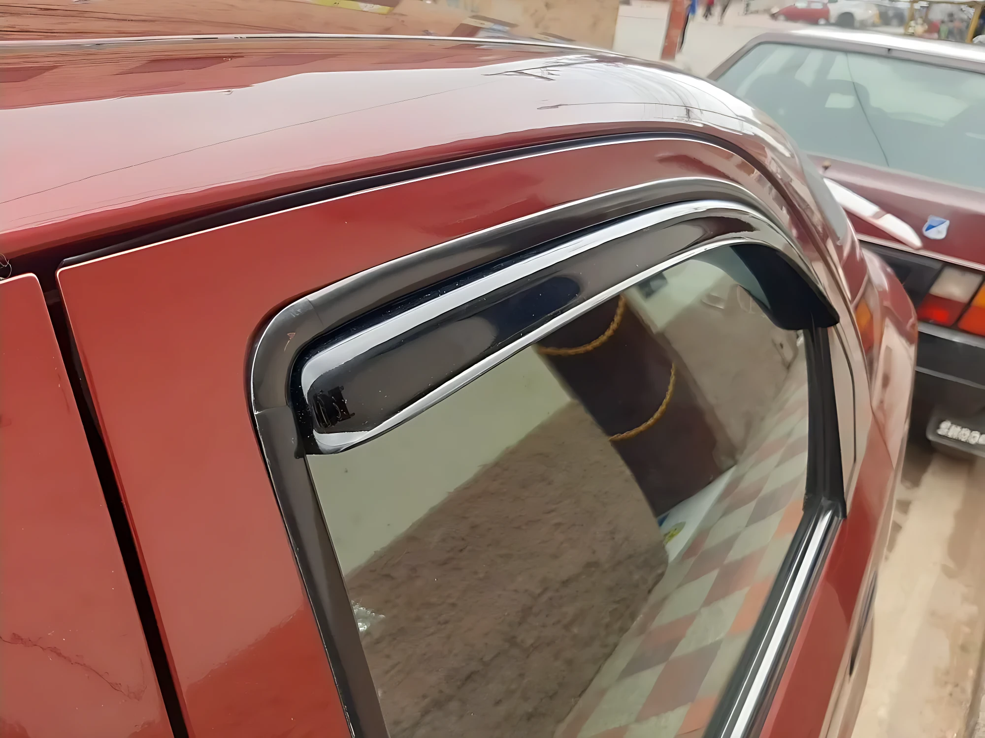 Deflector de ventana para auto en color rojo, instalado en la puerta lateral. Incluye goma flexible negra que sella el borde superior del cristal, permitiendo ventilación sin entrada de lluvia o viento.