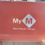 Caja roja con logo blanco que dice MyM y texto Micro Needle Therapy, sobre mostrador de vidrio en tienda.