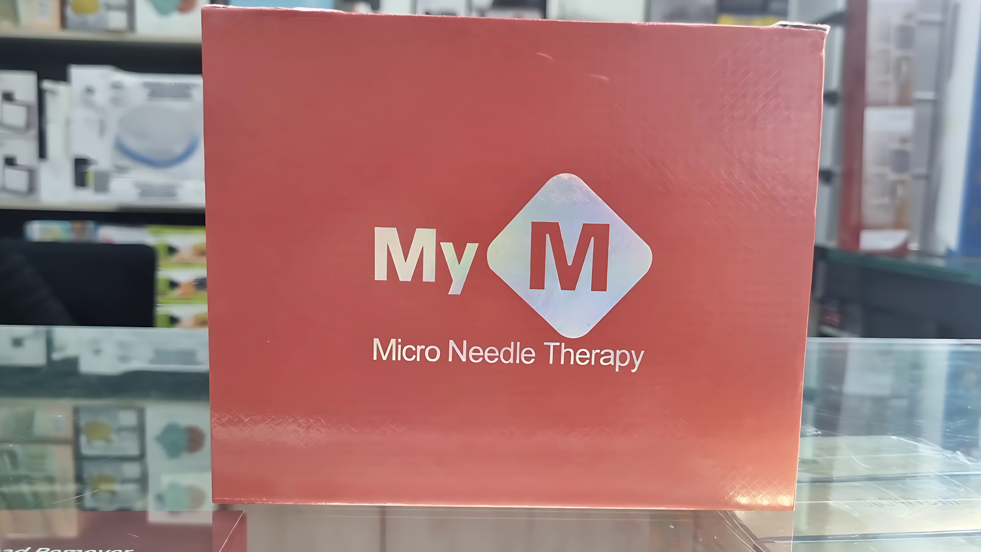 Caja roja con logo blanco que dice MyM y texto Micro Needle Therapy, sobre mostrador de vidrio en tienda.