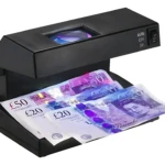 Detector de billetes falsos negro con luz UV y lupa, mostrando billetes de 50 y 20 libras bajo análisis en su bandeja.