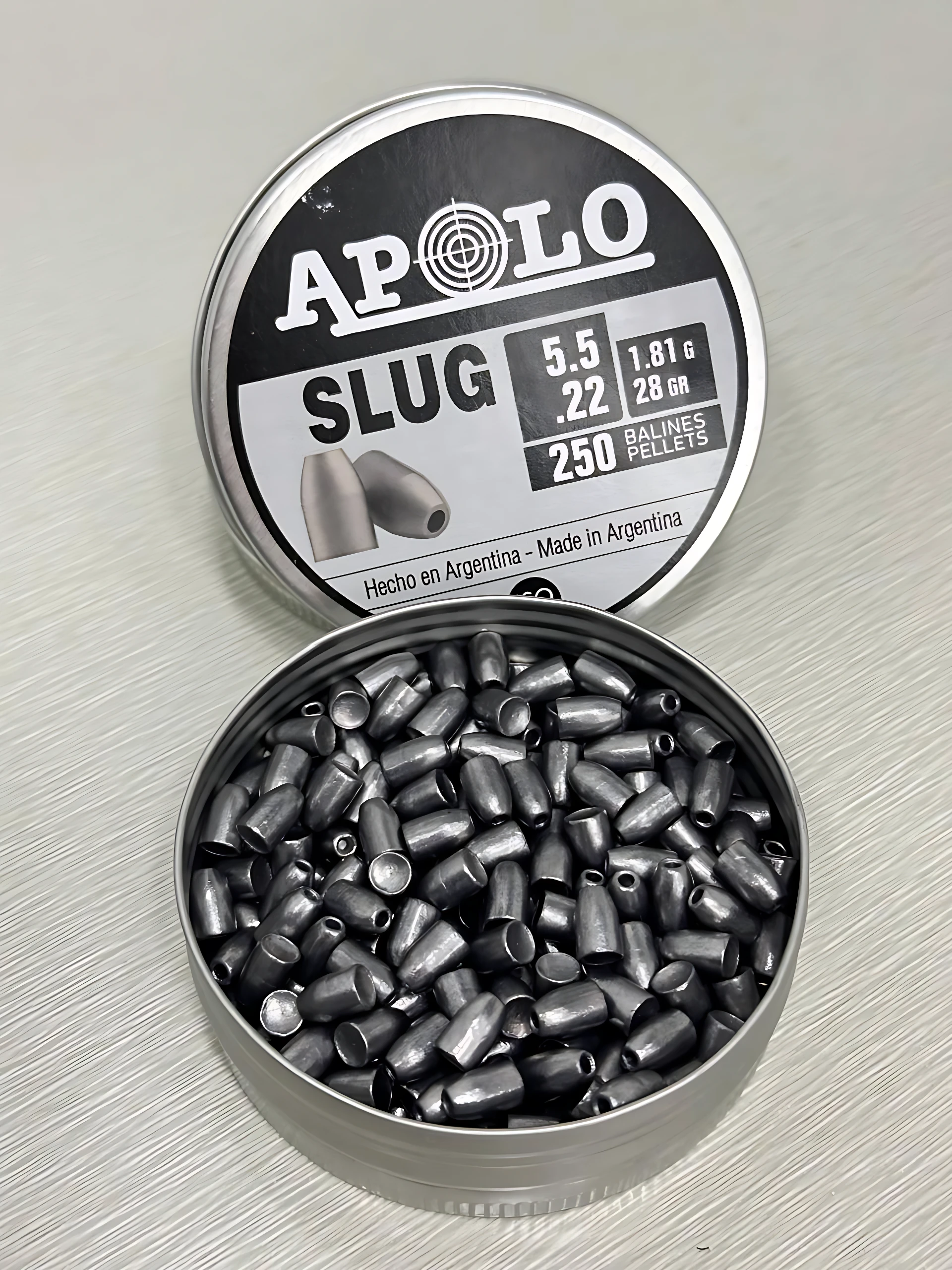 Lata abierta de perdigones Apolo Slug 5.5 mm calibre .22 de plomo color gris, mostrando su interior con balines dispuestos al azar y la tapa al fondo con especificaciones técnicas de 1.81 g y 28 gr.