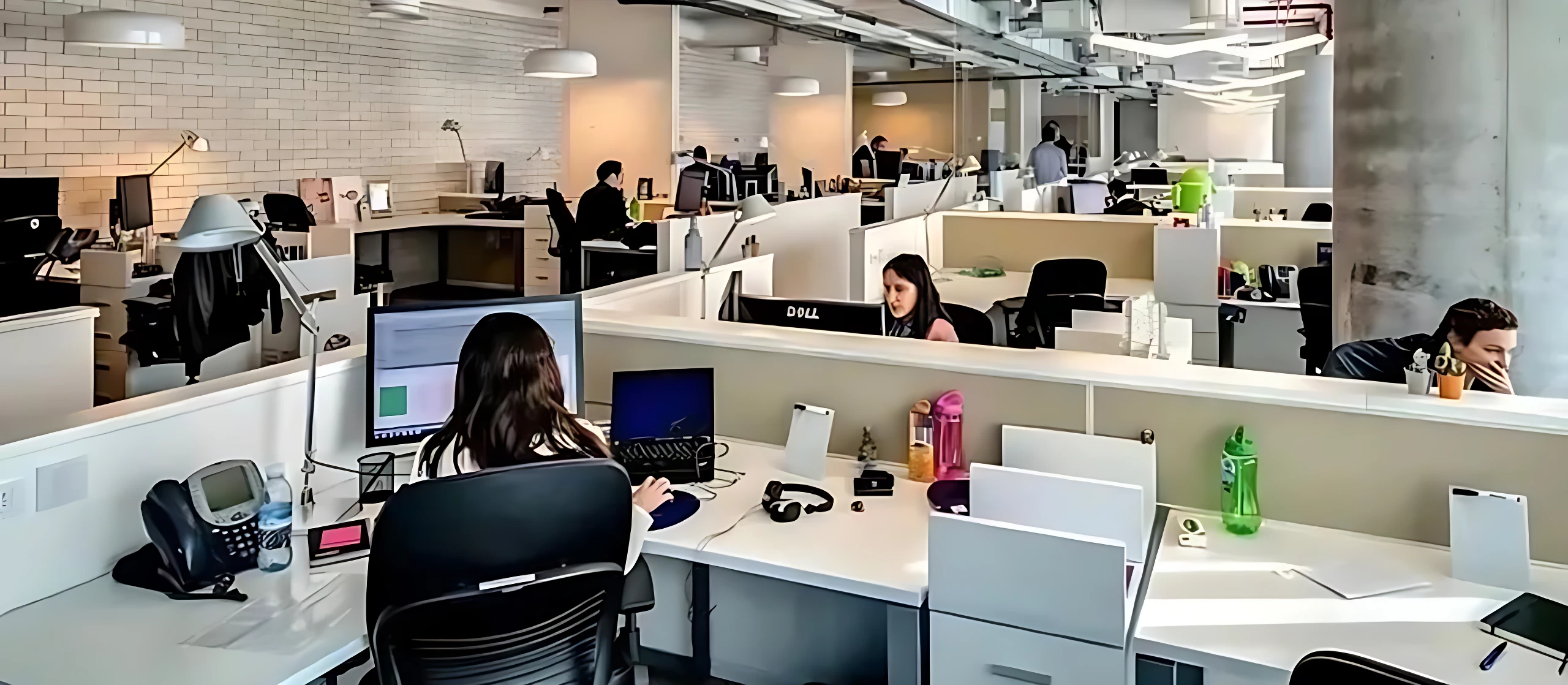Vista general de oficina abierta con empleados trabajando en escritorios equipados con computadoras, auriculares y accesorios. Ambiente profesional con iluminación natural y mobiliario moderno.