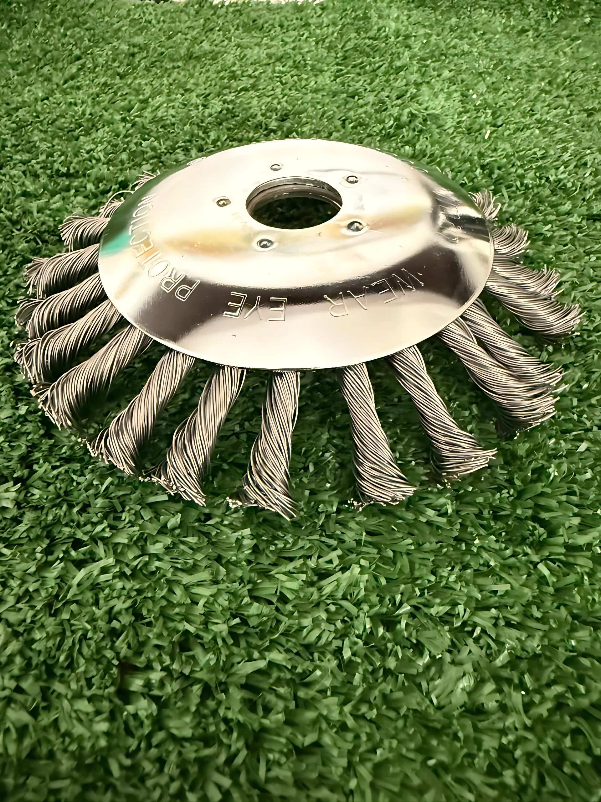 Disco de alambre trenzado con base metálica plateada, diseñado para eliminar malezas y óxido. Visto desde arriba sobre fondo verde artificial.