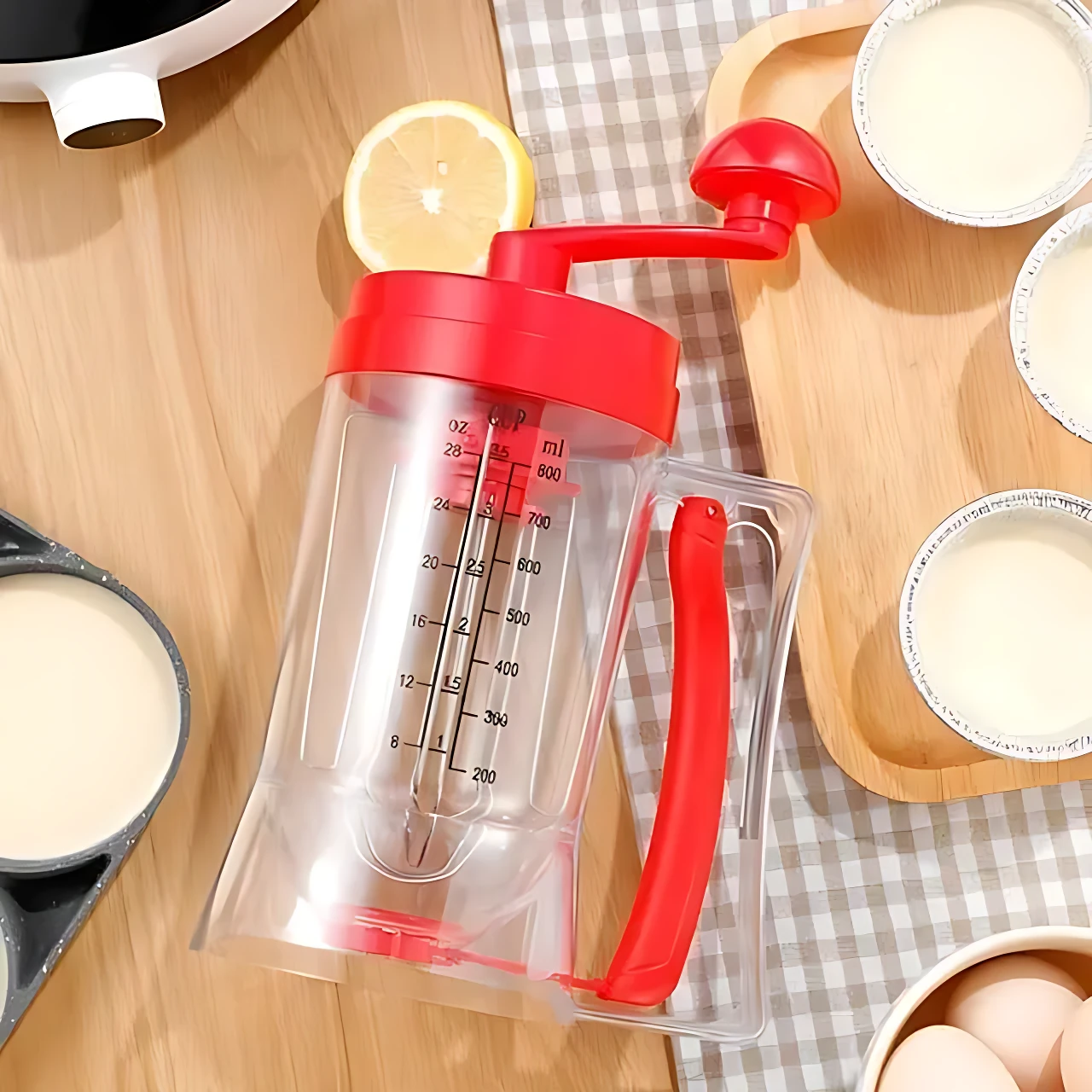Dispensador de masa con mango rojo y medidor interno, sobre mesa de madera con limón y tazas de leche al fondo.