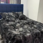 Edredón térmico peludo negro sobre cama doble con cabecero azul tapizado, textura suave y voluminosa en ambiente interior.