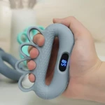 Ejercitador de manos azul claro con pantalla digital mostrando número 7, textura rugosa en agarre y tres anillos para dedos, sostenido por mano derecha.