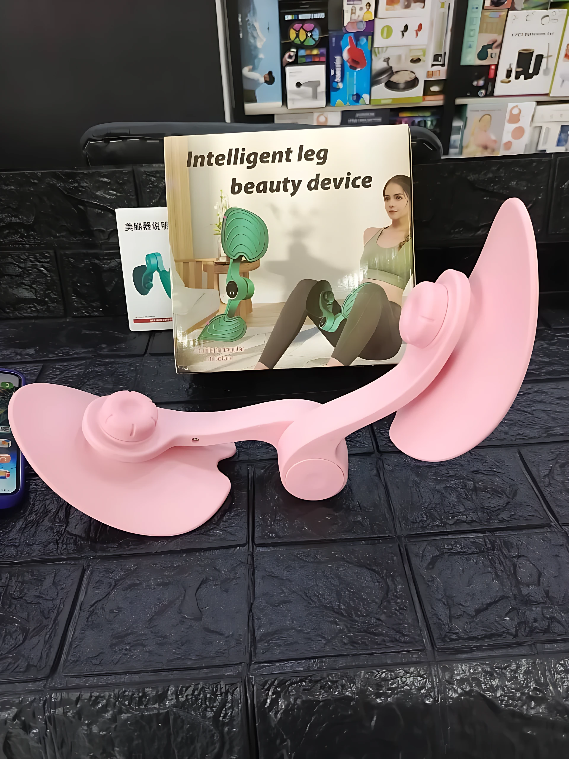 Dispositivo rosa para ejercicio de piernas y cadera, con base plana y soportes ergonómicos, acompañado de su caja que muestra de uso.