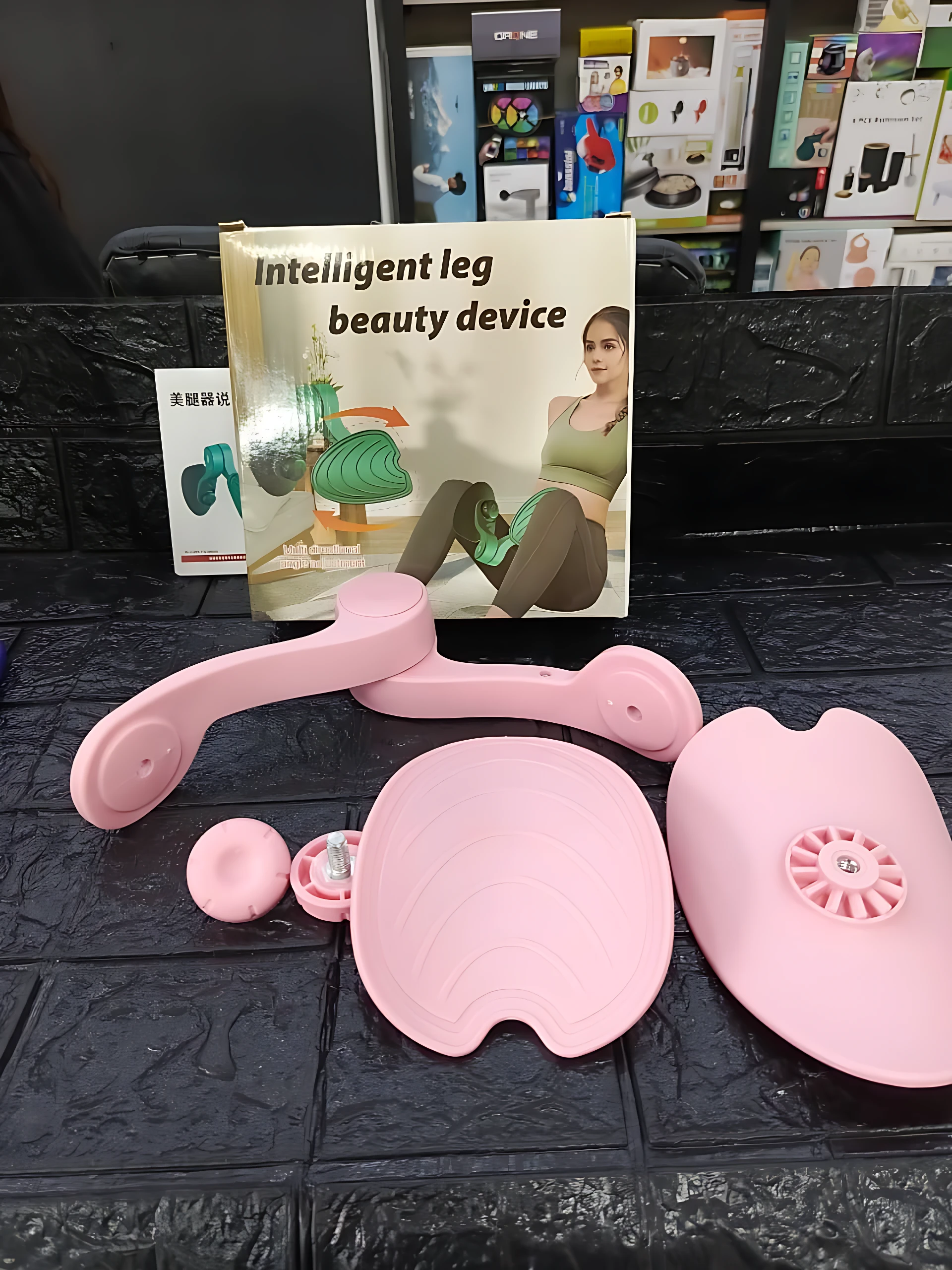 Dispositivo ergonómico para tonificar piernas y cadera, color rosa. Incluye soporte con mango, almohadillas y accesorios. Presentado junto a su caja.