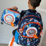 Niño de espaldas con mochila escolar azul espacial y lonchera térmica, ambos con estampado de astronautas y detalles naranjas.