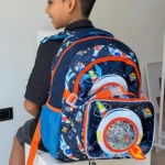 Morral escolar infantil azul espacial con lonchera térmica integrada, usado por niño sentado en caja blanca.