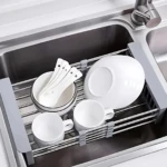 Escurridor de acero inoxidable con rejilla, en uso dentro de un fregadero, secando platos, tazas y cubiertos blancos.