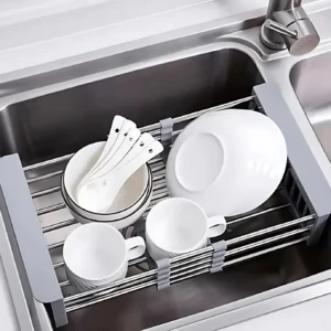 Escurridor de acero inoxidable con rejilla, en uso dentro de un fregadero, secando platos, tazas y cubiertos blancos.
