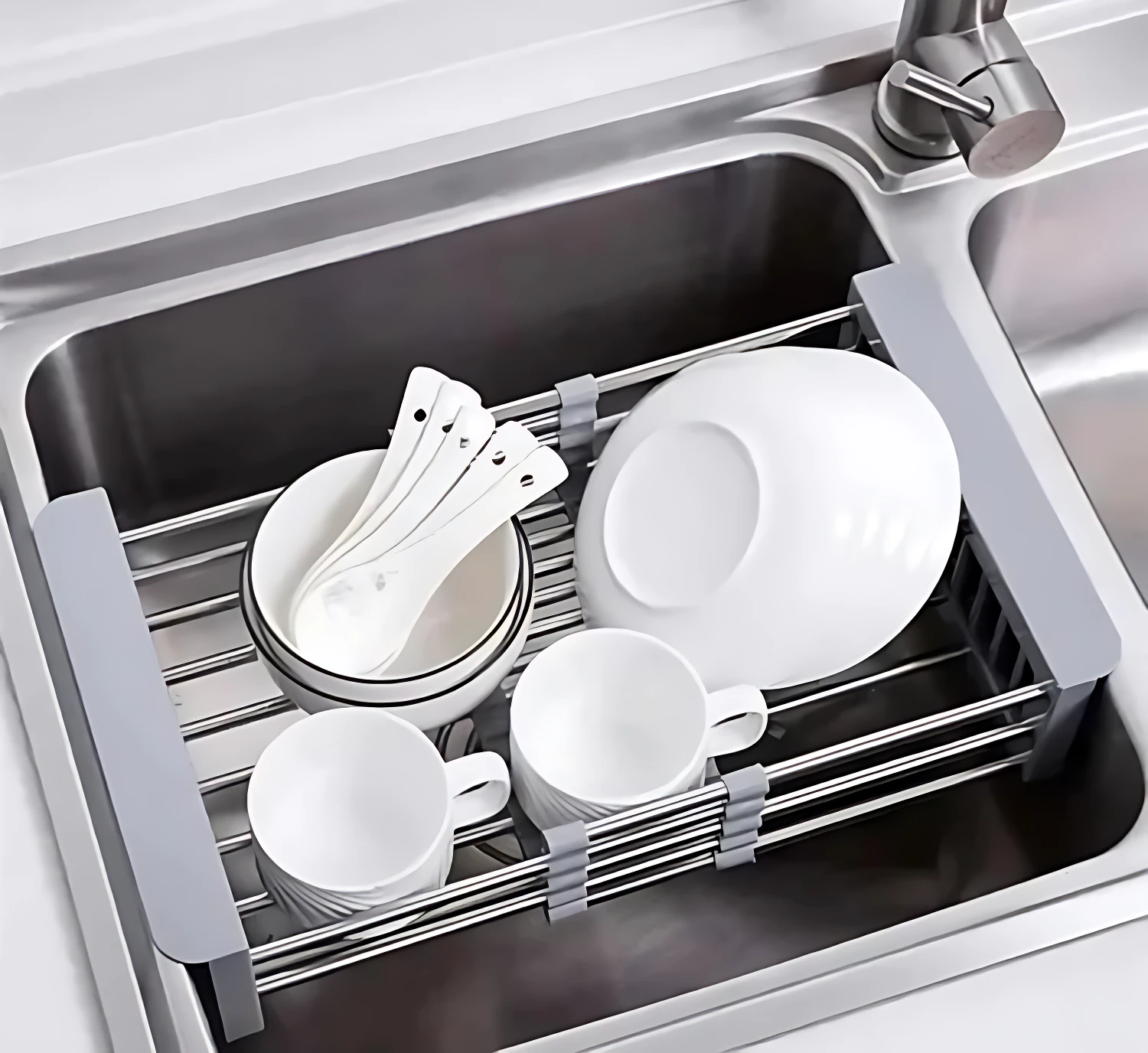 Escurridor de acero inoxidable con rejilla, en uso dentro de un fregadero, secando platos, tazas y cubiertos blancos.