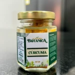 Jarrón hexagonal de vidrio con tapa dorada, etiqueta blanca y verde que indica "Cúrcuma" y "50 GR", sobre superficie oscura brillante.