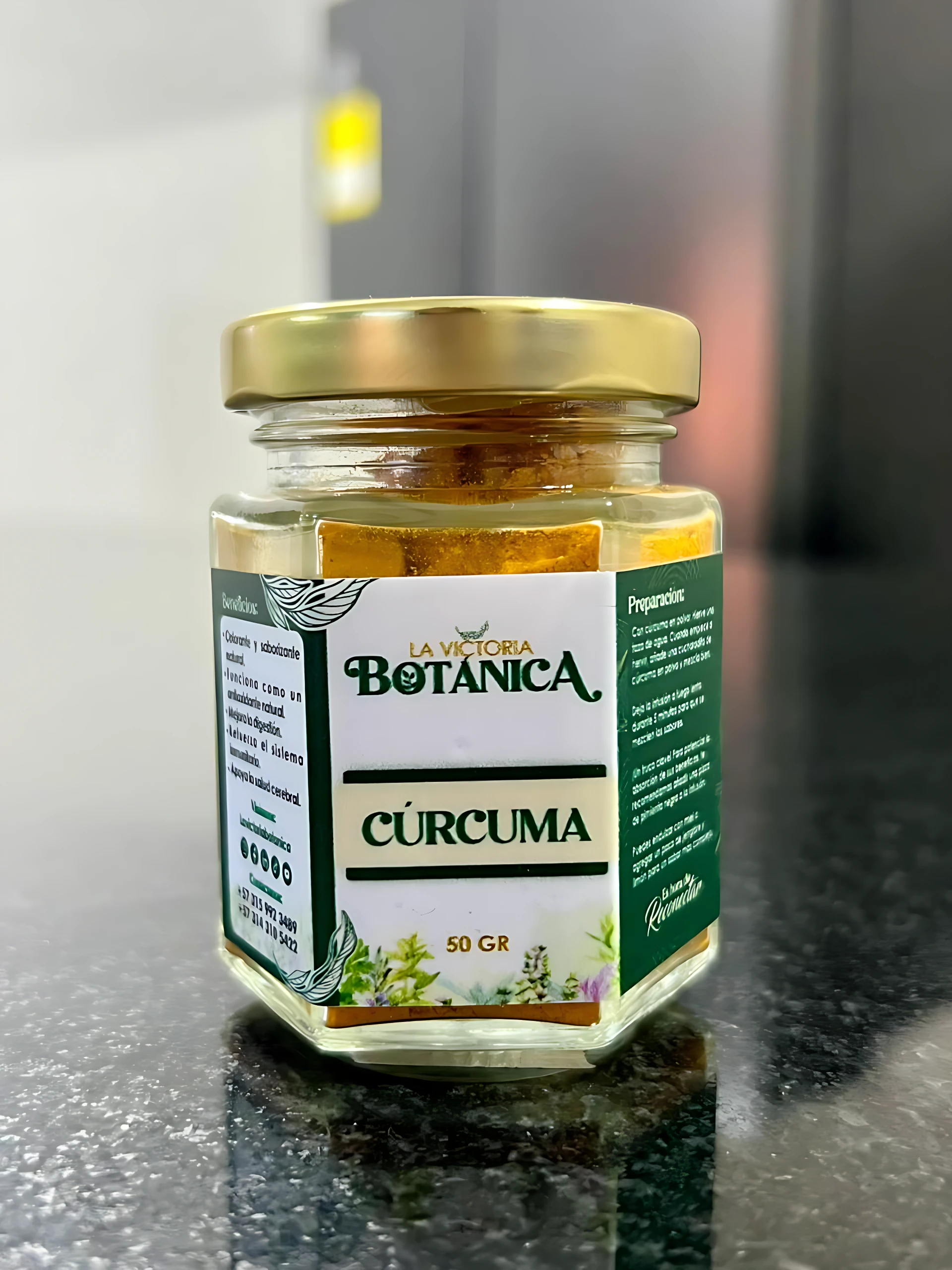 Jarrón hexagonal de vidrio con tapa dorada, etiqueta blanca y verde que indica "Cúrcuma" y "50 GR", sobre superficie oscura brillante.