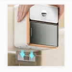 Dispensador de papel higiénico blanco con cajón superior y estante inferior, montado en pared beige con adhesivo transparente visible.
