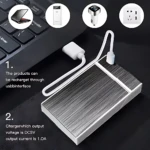 Estuche rectangular metálico con panel solar y cable USB, sobre teclado negro. Incluye iconos de carga por USB, encendedor y enchufe. Texto indica recarga vía USB y salida de 1.0A.