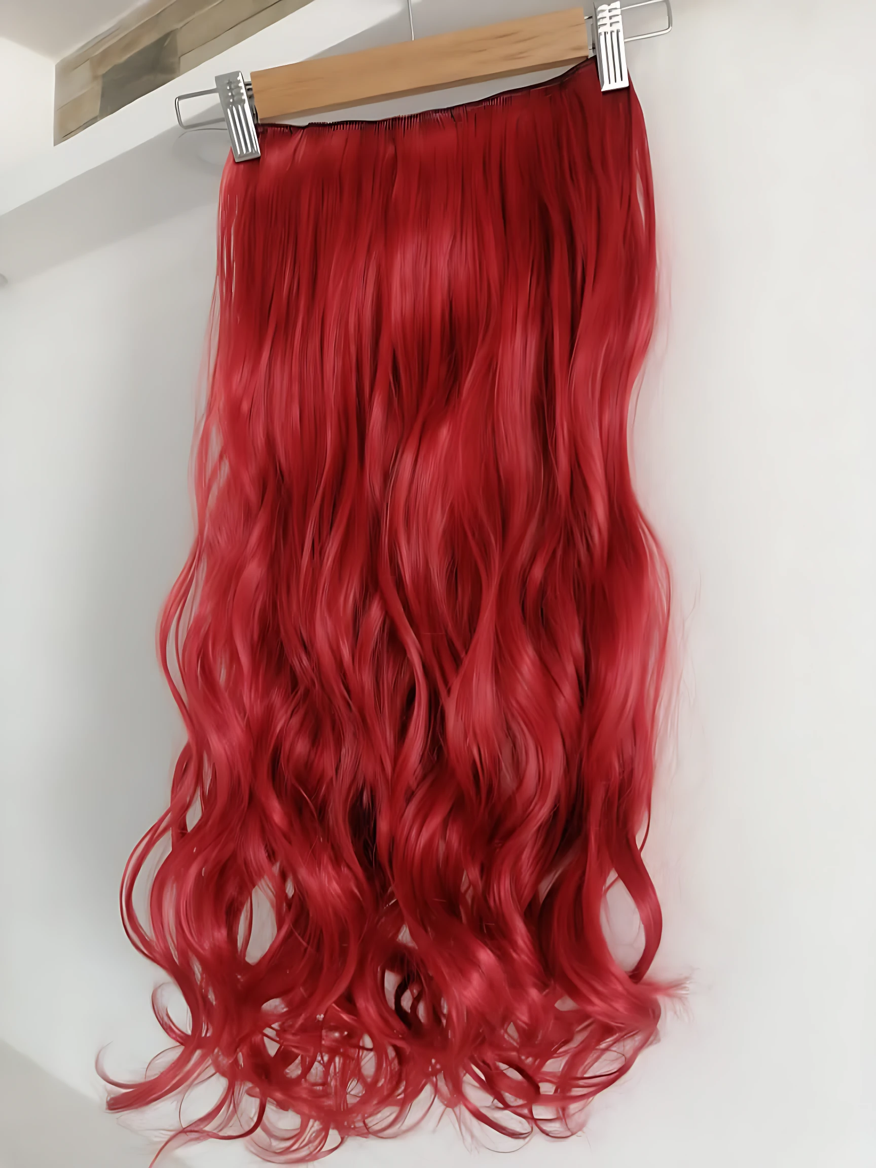 Extensiones semi humanas onduladas rojas con clips metálicos, colgadas en perchero de madera sobre fondo blanco.