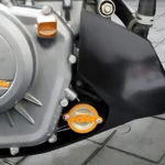 Tapa filtro aceite KTM acero inoxidable, acabado brillante con logo naranja, instalada en motor de motocicleta sobre suelo de baldosas grises.