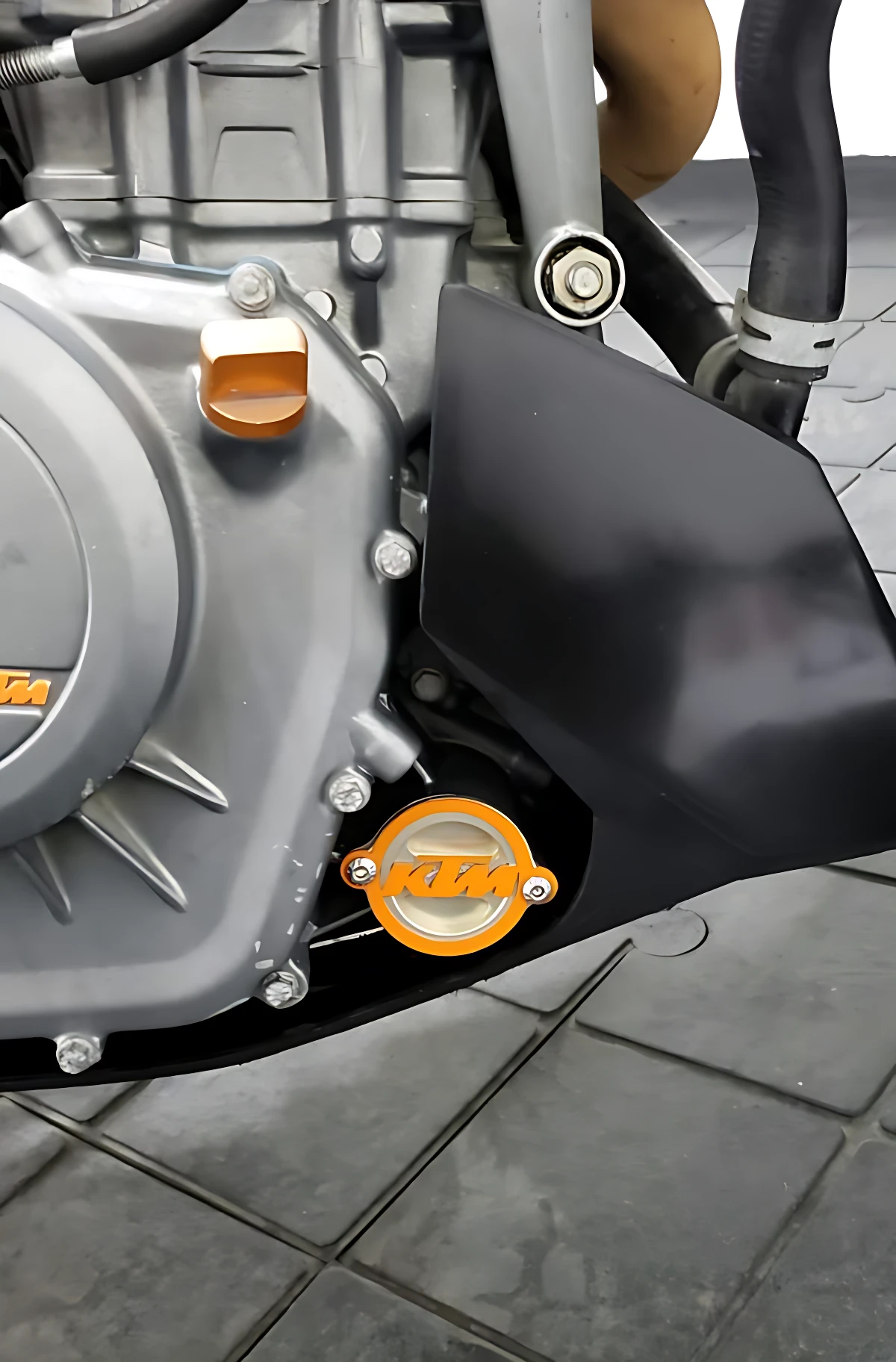 Tapa filtro aceite KTM acero inoxidable, acabado brillante con logo naranja, instalada en motor de motocicleta sobre suelo de baldosas grises.