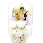 Domo de vidrio con arreglo floral eterno en blanco y amarillo, sobre base negra circular. Incluye rosa blanca, flores amarillas y hojas verdes, con detalles de cintas blancas. Medidas visibles: 21 cm de altura por 11 cm de ancho.