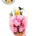 Domo de vidrio con arreglo floral eterno de rosas rosadas, flores amarillas y hojas verdes sobre base negra. Medidas 21 cm de alto por 11 cm de ancho.