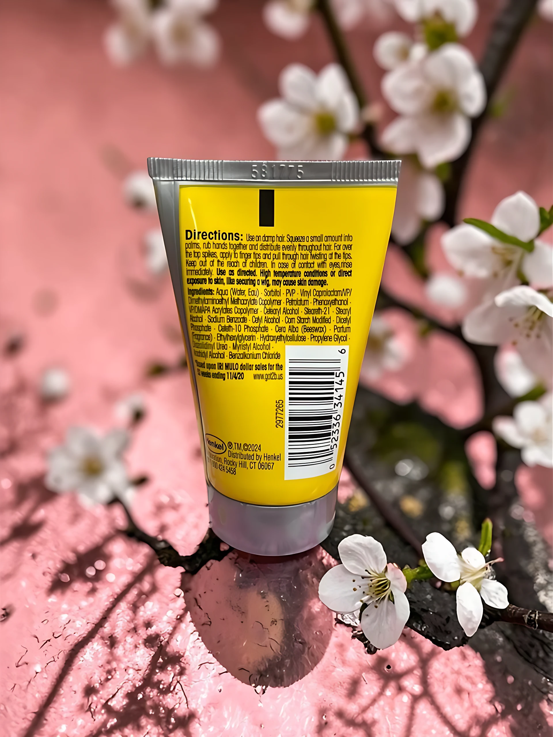 Tubo amarillo de gel para cabello Got2b con instrucciones visibles, sobre rama con flores blancas.