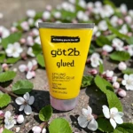 Tubo amarillo de gel para cabello Got2b Glued, con texto negro y detalles en blanco, sobre fondo de flores blancas y verdes.