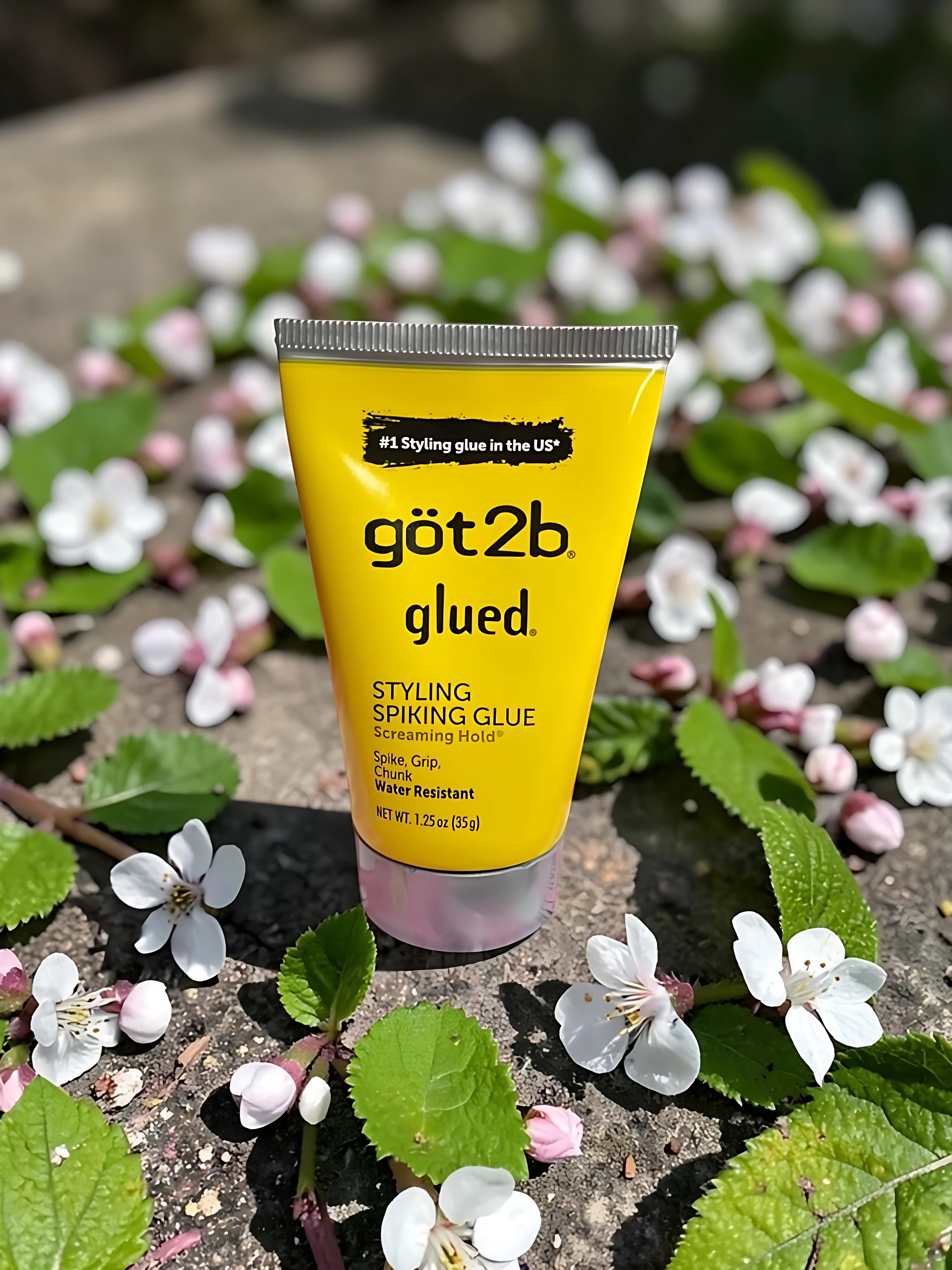 Tubo amarillo de gel para cabello Got2b Glued, con texto negro y detalles en blanco, sobre fondo de flores blancas y verdes.