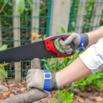Guante de seguridad en malla de acero inoxidable con refuerzo azul, usado mientras se corta madera con sierra manual en jardín.