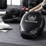 Guantes de boxeo Wonder en cuero PU negro, con diseño ergonómico y protección para muñeca, sobre superficie gris con accesorios de entrenamiento al fondo.