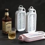 Dos hieleras portátiles blancas con asas, una abierta mostrando hielo y otra cerrada, junto a una tercera en color rosa abierta, acompañadas de una botella de Jack Daniel’s y un vaso con bebida.