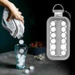 Hielera portátil en uso: mano vierte bolas de hielo de un molde flexible blanco a vaso transparente sobre mesa oscura con frutas al fondo.