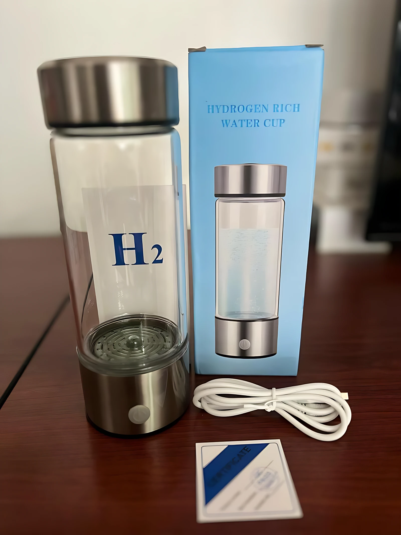 Ionizador de agua inteligente con hidrógeno molecular, tetera transparente con tapa metálica, base plateada y cable blanco incluido, sobre mesa de madera.