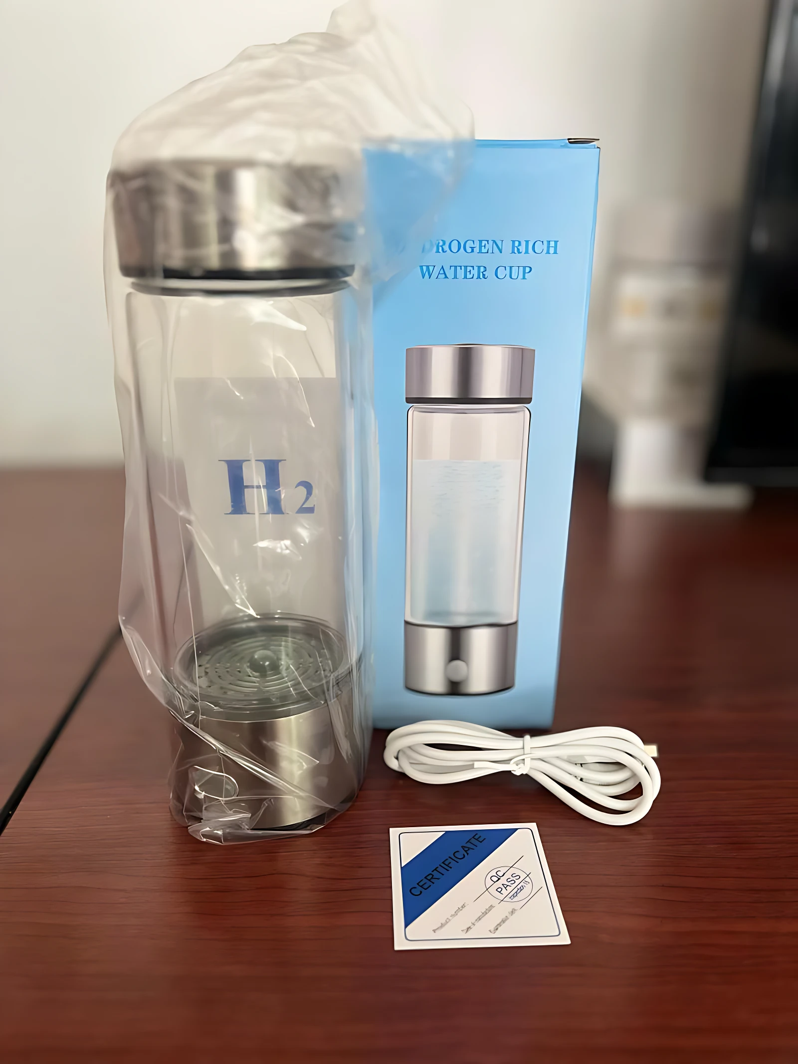 Ionizador de agua inteligente con hidrógeno molecular, envuelto en plástico transparente, junto a su caja azul y cable de carga sobre mesa de madera.