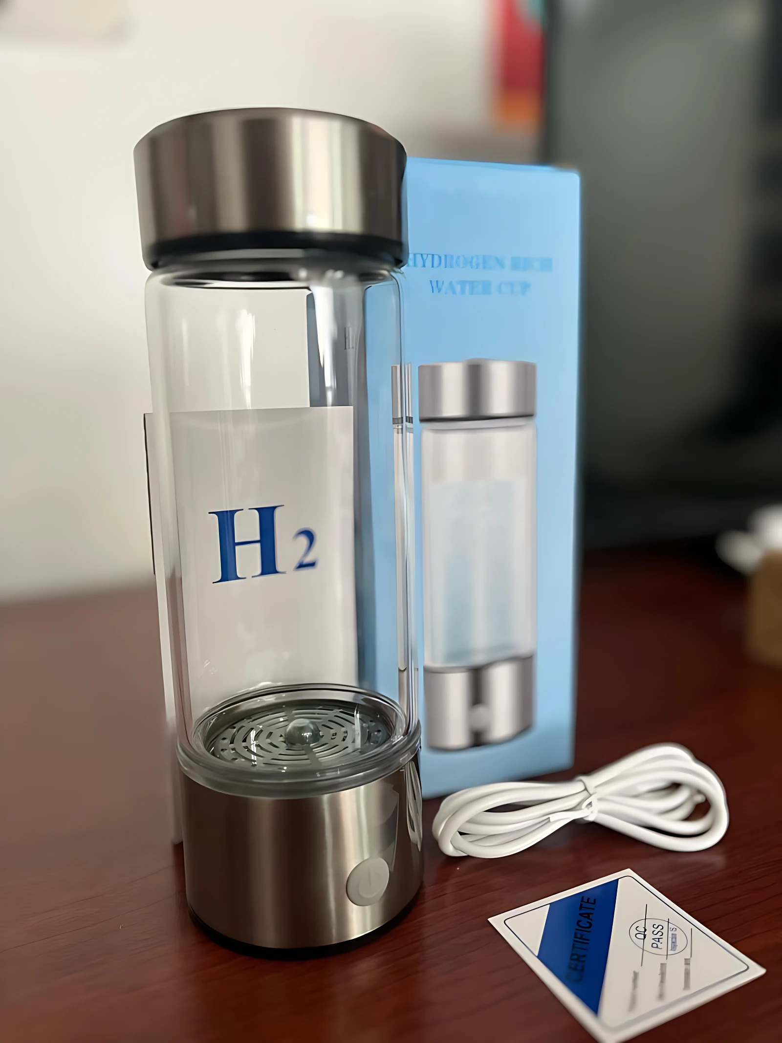 Ionizador de agua inteligente con hidrógeno molecular, cuerpo de vidrio transparente y base metálica, acompañado de su caja azul, cable de carga y tarjeta de garantía sobre superficie oscura.