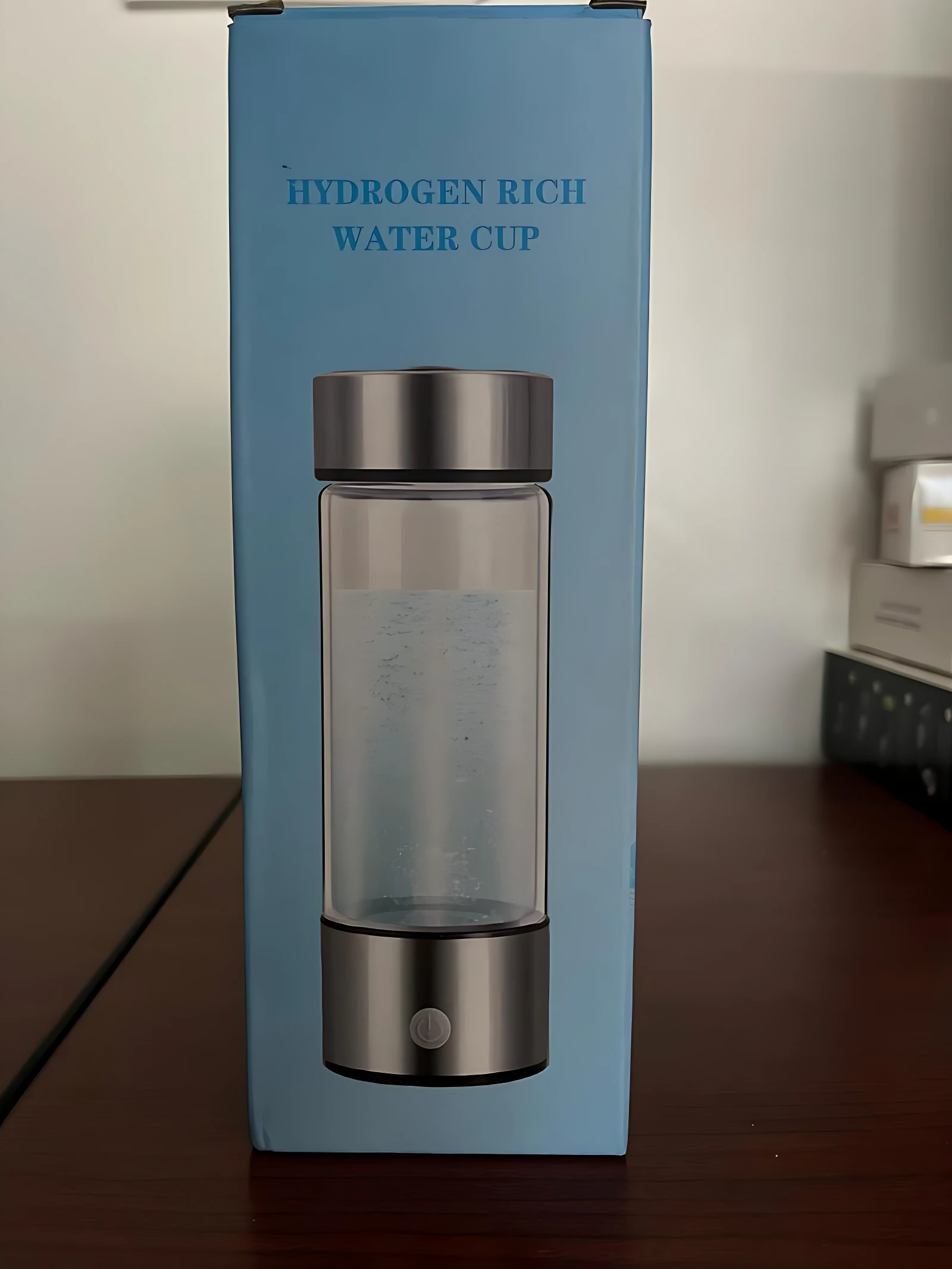 Caja azul del ionizador de agua inteligente con hidrógeno molecular, mostrando el vaso transparente con tapa y base metálicas sobre mesa de madera.