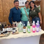 Tres personas posan frente a una mesa con jabones corporales naturales Zoieco en botellas de vidrio transparente con etiquetas blancas y tapas de colores, sobre mantel blanco con borla.